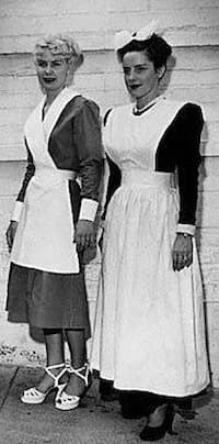 This is a black and white photo showing two Harvey Girl uniforms. On the right is the classic one with a long black dress and full white bib apron. On the left is a woman in a slightly shorter black dress with white cuffs. The apron has a criss-cross bib top, and she is wearing white sandals instead of black shoes.