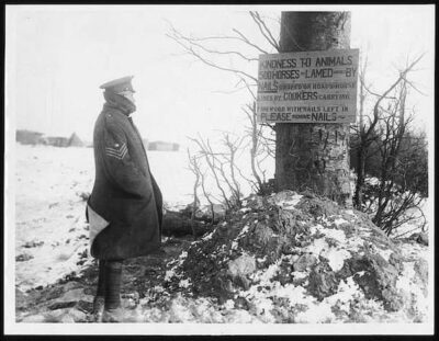 A photo of a military officer reading a sign that reminds people to remove nails from wood they are using. Otherwise they harm the horses.