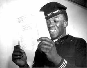This photo is of a Black member of the Coast Guard. He is smiling and enjoying reading a letter. 