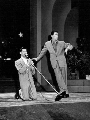 Dean Martin leans comfortably on a piece of furniture while Jerry Lewis is on his knees holding a microphone. 
