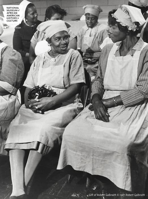 This is a black-and-white photo of two midwives in uniform sitting and talking or waiting for a class to begin. Other midwives are in the background.