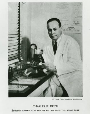A black-and-white photo of Dr. Charles Drew in a medical laboratory, sitting in frong of a microphone.