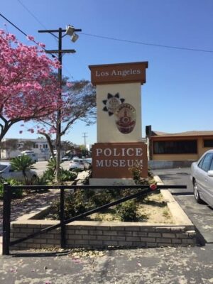 This color photo shows the Los Angeles Police Museum sign. There is a blooming cherry blossom tree to the left of it.