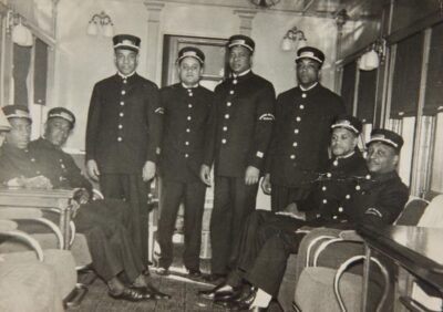Black-and-white photo of the Sleeping Car Porters in a train car. They are all in uniform, wearing hats.