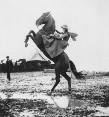 Annie Oakely in a dress and hat rides a rearing horse on muddy ground, with a tent and several people in the background.