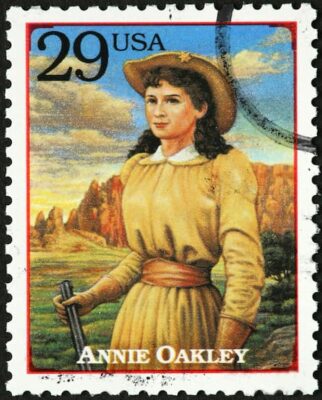 U.S. postage stamp honoring Annie Oakley. A blue sky and mountainous red rocks are behind her. She wears a hat and is dressed in yellow.