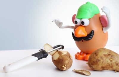 A more recent Mr. Potato Head is pictured in sneakers with a baseball cap on. He watches in surprise as a potato peeler peels a regular potato.