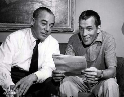 Richard Rodgers wears a dress shirt and tie. Ed Sullivan is in a sport shirt. He is holding a script or an article that he seems to be reviewing with Rodgers.