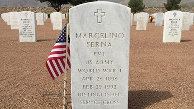 A gravestone for Marcelino Serna, U.S. Army, World War I, with an American flag beside it honors the decorated soldier. Other gravestones and trees are visible in the background, with mountains in the distance.