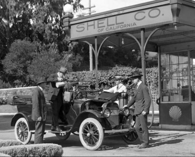 This is a black-and-white photo. Two businessmen and a Shell Company employee stand by the automobile. The driver is adding the gasoline.