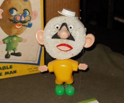 Mr. Potato Head with a Styrofoam head stands in front of a boxed set of Mr. Potato Head. He wears a white boater hat, a yellow suit, and has on green shoes/feet.