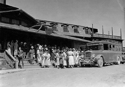 This is a black-and-white photograph of tourists dressed in suits and dresses (1930s length skirts). They are lined up outside El Tovar, the elegant hotel along the Grand Canyon rim. The photo was originally from the Fred Harvey Collection.