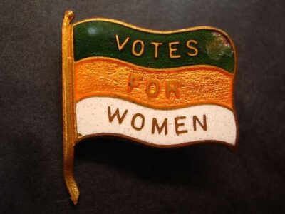 A pin shaped like a waving flag with green, orange, and white stripes, reading VOTES FOR WOMEN in bold letters—a powerful symbol of women participation at every national political convention.