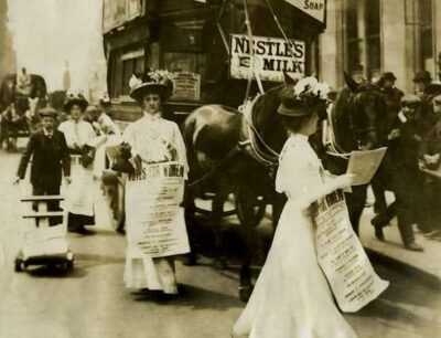 Women in early 1900s dresses and hats march in a street protest, holding papers and banners about voting rights, marking a significant moment for Women Participation. A horse-drawn carriage and onlookers are visible in the background, underscoring the historic push that would soon lead to Firsts for Women at events like the National Political Convention.