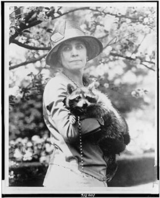 Grace Coolidge in a hat and suit holding Rebecca the raccoon for aphotograph.
