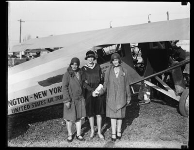 Three women standi n their hats and coats, likely awaiting instructions to board what is a rather primitive airplane.